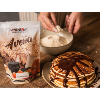 Harina de avena Elevenfit Conguix 1kg junto a un postre de panqueques con chocolate, en fondo de mesa.